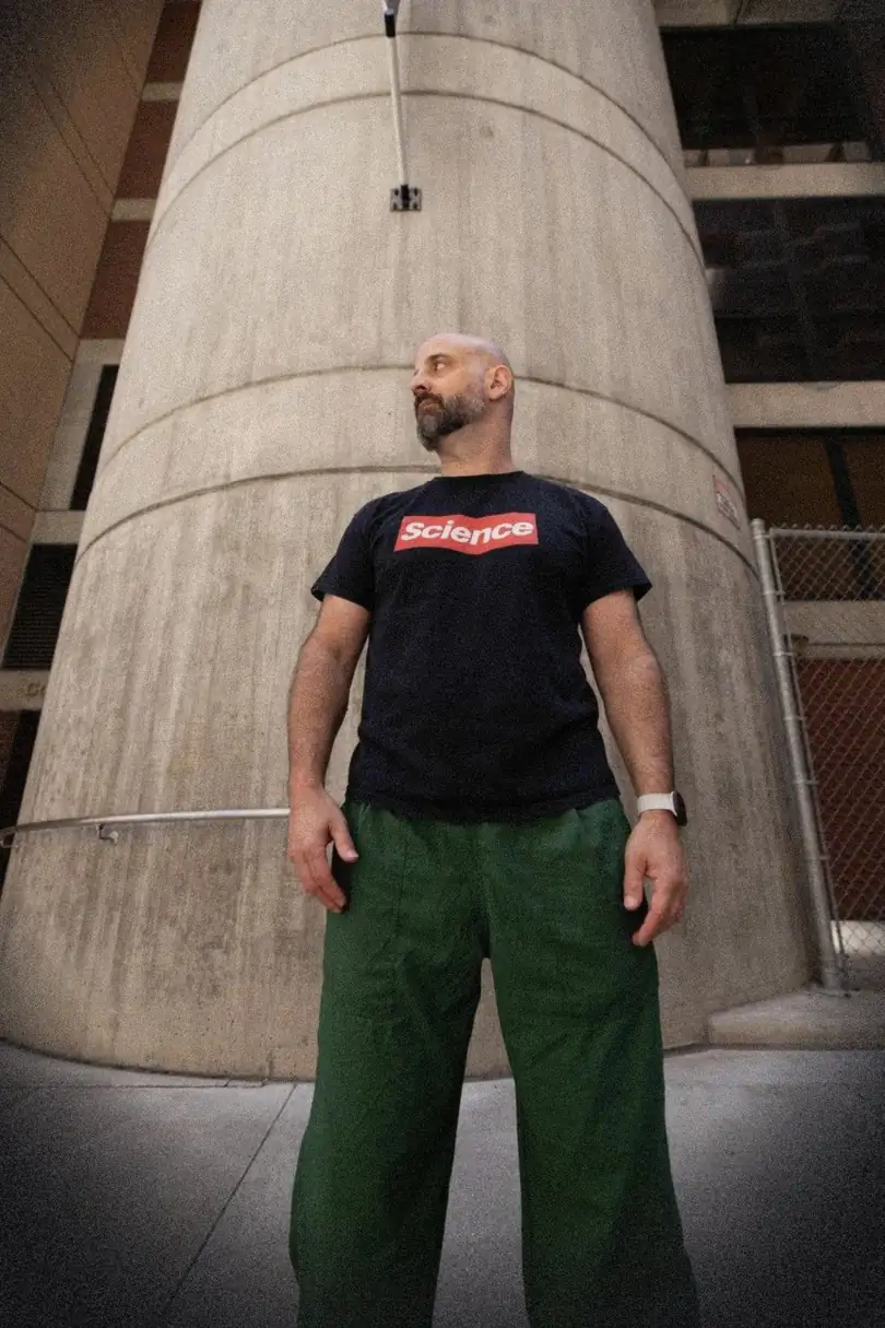 MING outdoors in a black Science t-shirt and green pants, standing in front of a concrete column